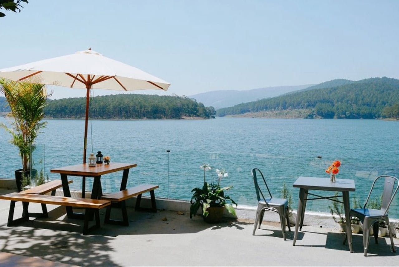 about-01 Enjoy a serene lakeside dining experience with wooden tables under a sun umbrella.