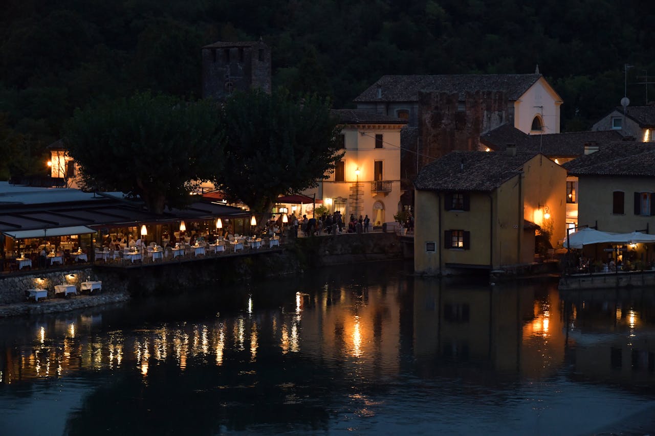 services-03 Illuminated waterfront restaurants and reflections at dusk in Borghetto, Veneto, Italy.